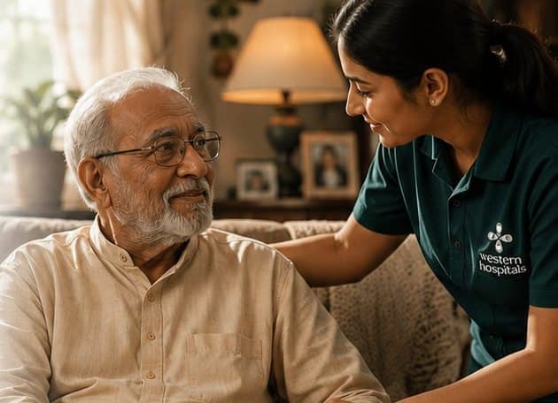 A Western Hospitals care monitor sits beside an elderly man in his living room, a hand on his shoulder, talking with him.